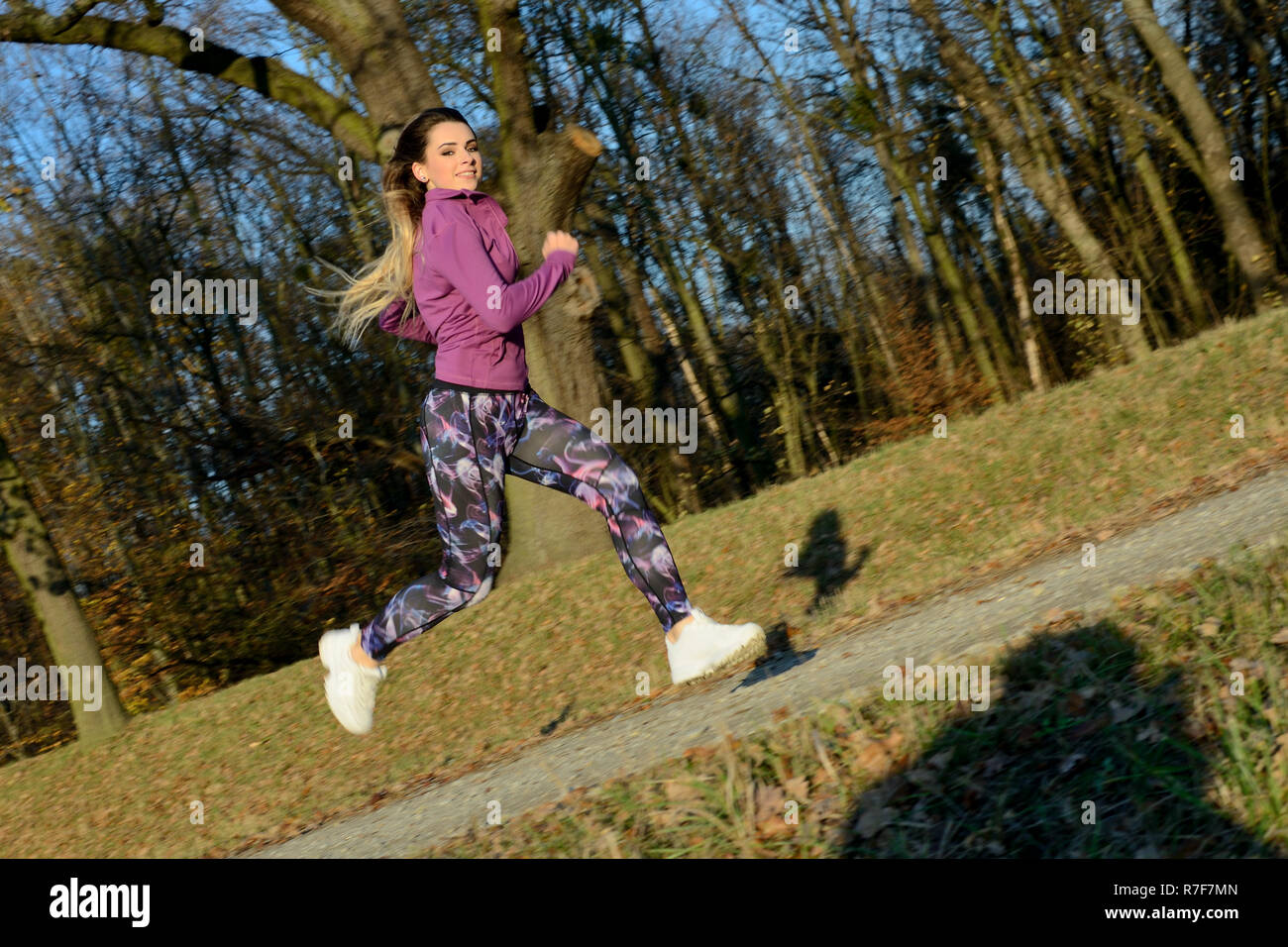 Young girl running in the forest. Blonde female with colorful leggings and purple top. Active runner with released hairs. Stock Photo