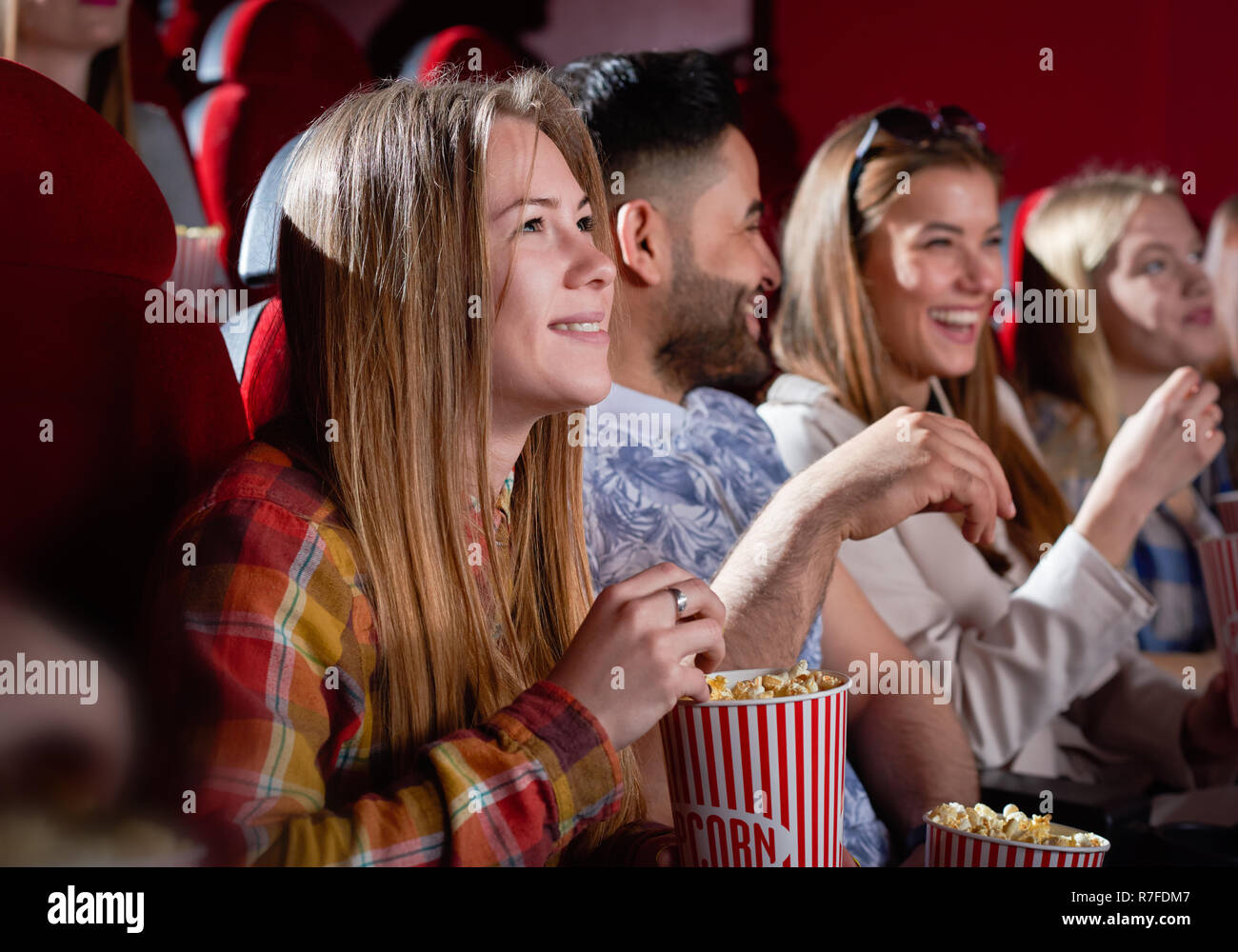 Smiling beautiful woman watching movie in cinema hall. Frightened attractive blonde wearing red checkered shirt keeping popcorn. Concept of entertainment and movie time.  Stock Photo