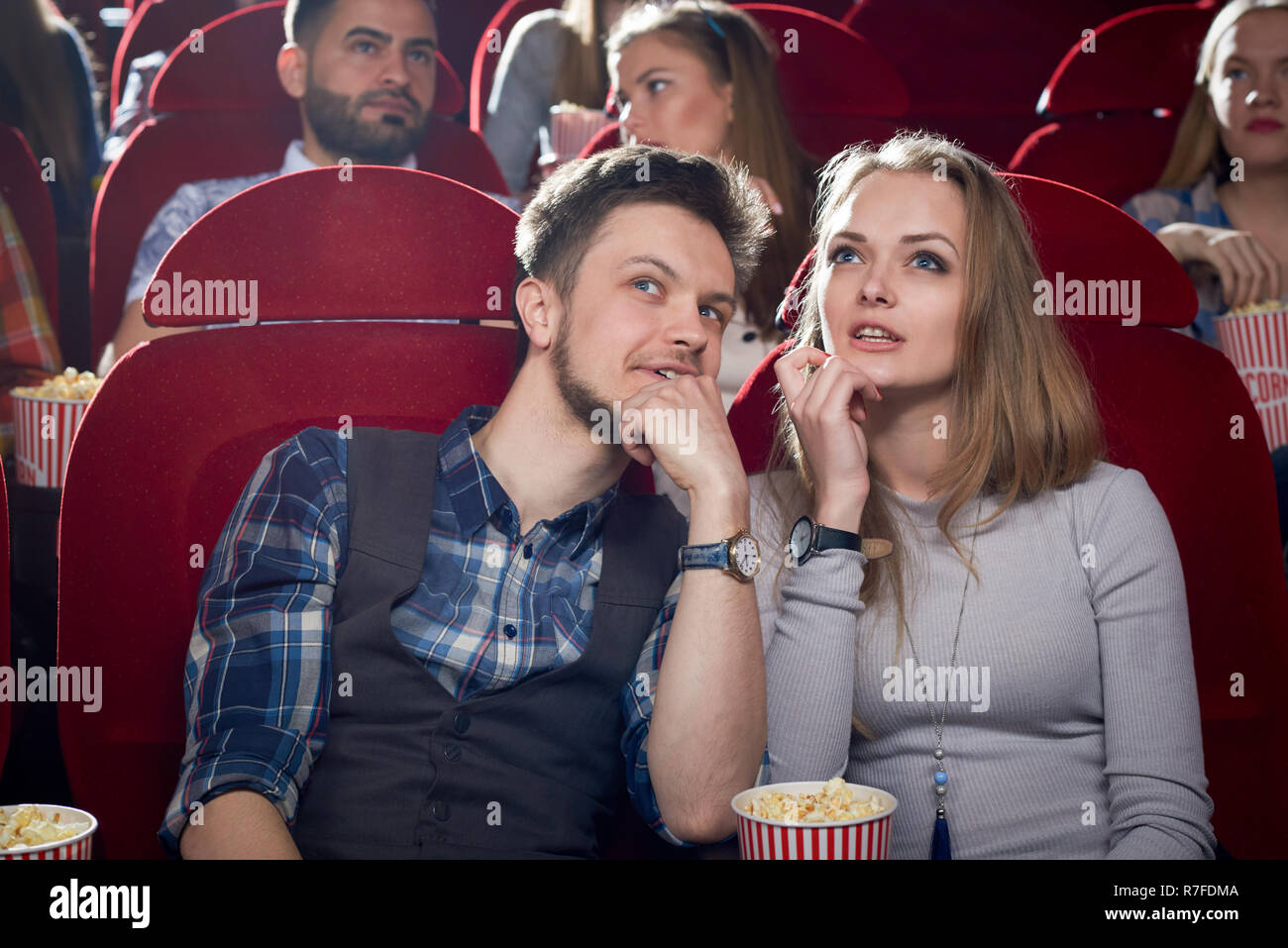 Cheerful couple having date in cinema. Smiling man talking it beautiful blonde girlfriend in gray. Cute pair watching interesting comedy and enjoying film together. Concept of free time of couple. Stock Photo