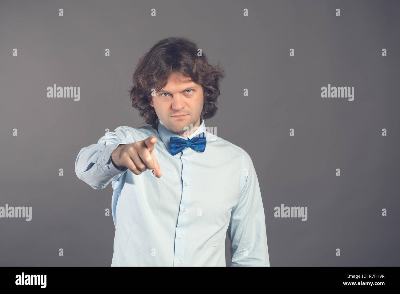 Young man in blue shirt with bow tie with shaggy hair, finger pointing directly towards the camera with index finger, blames you in doing wrong things on a gray background. Hey you. Studio shot. Stock Photo