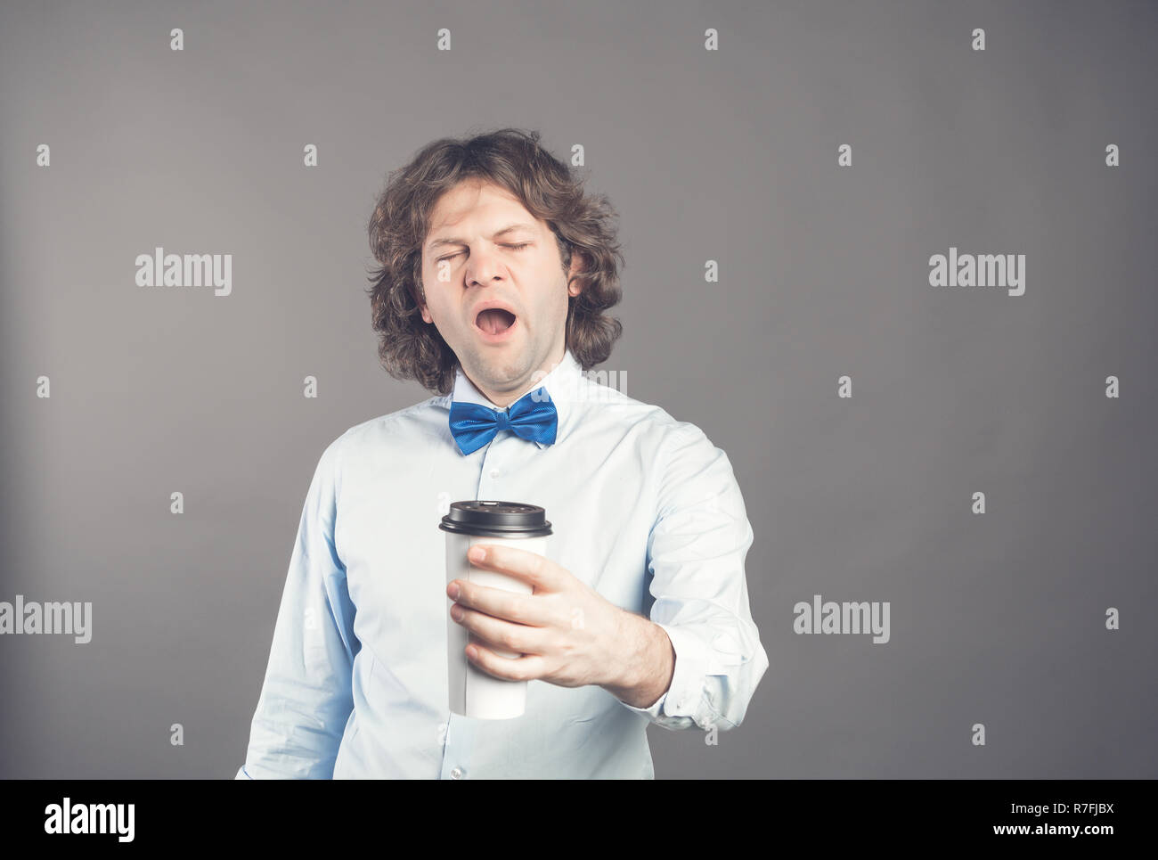 Tired guy yawns with sleepy expression and wide open mouth, holds disposable paper cup of coffee, dressed in shirt and blue bow tie poses against grey background. Morning time. Coffee break concept Stock Photo