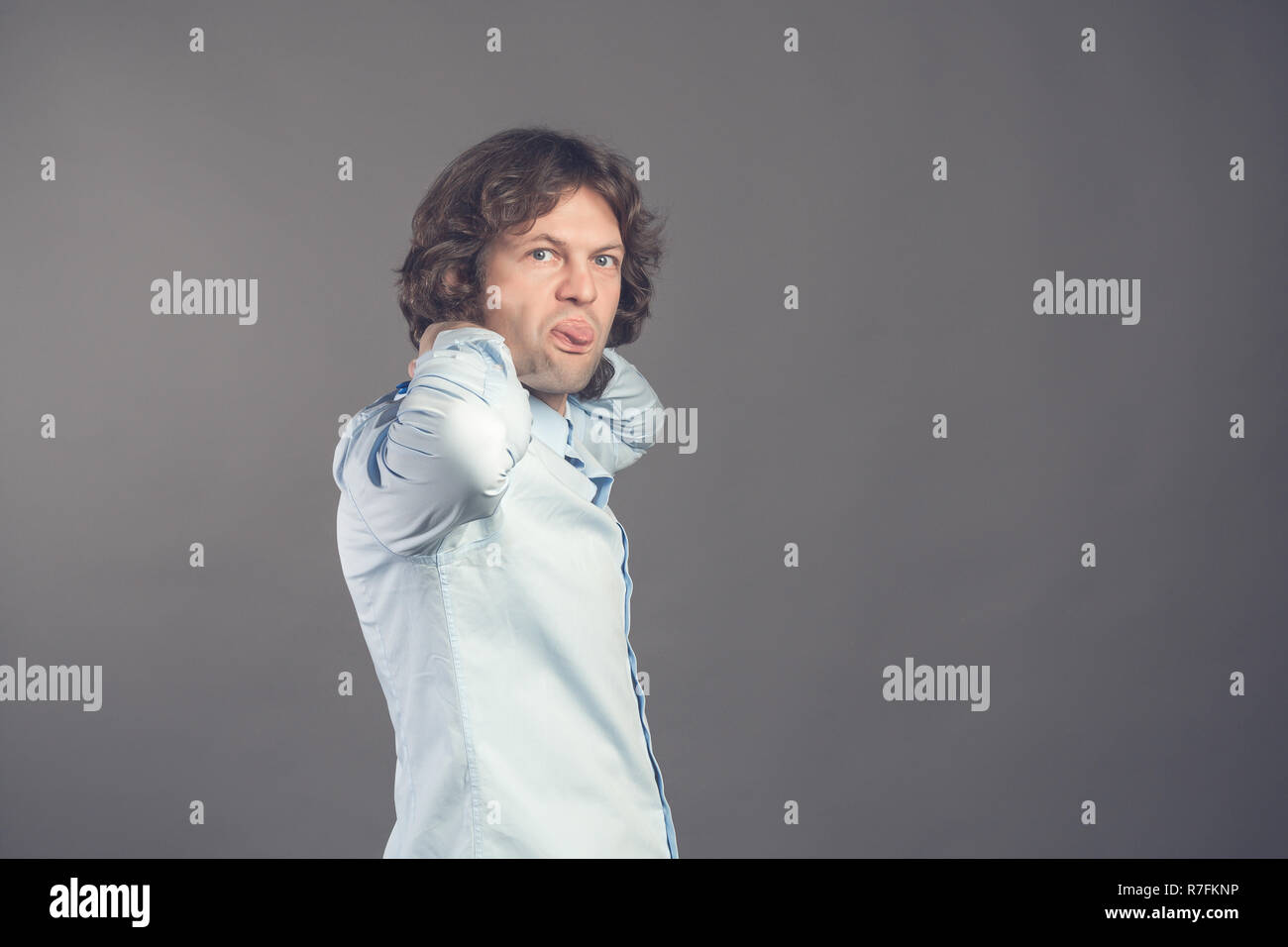 Man in shirt and bow tie with wavy hair on a grey background smiles and shows humorously the tongue. Standing half-turned male stuck his tongue right into the camera. Horizontal. Isolated. Copy space Stock Photo