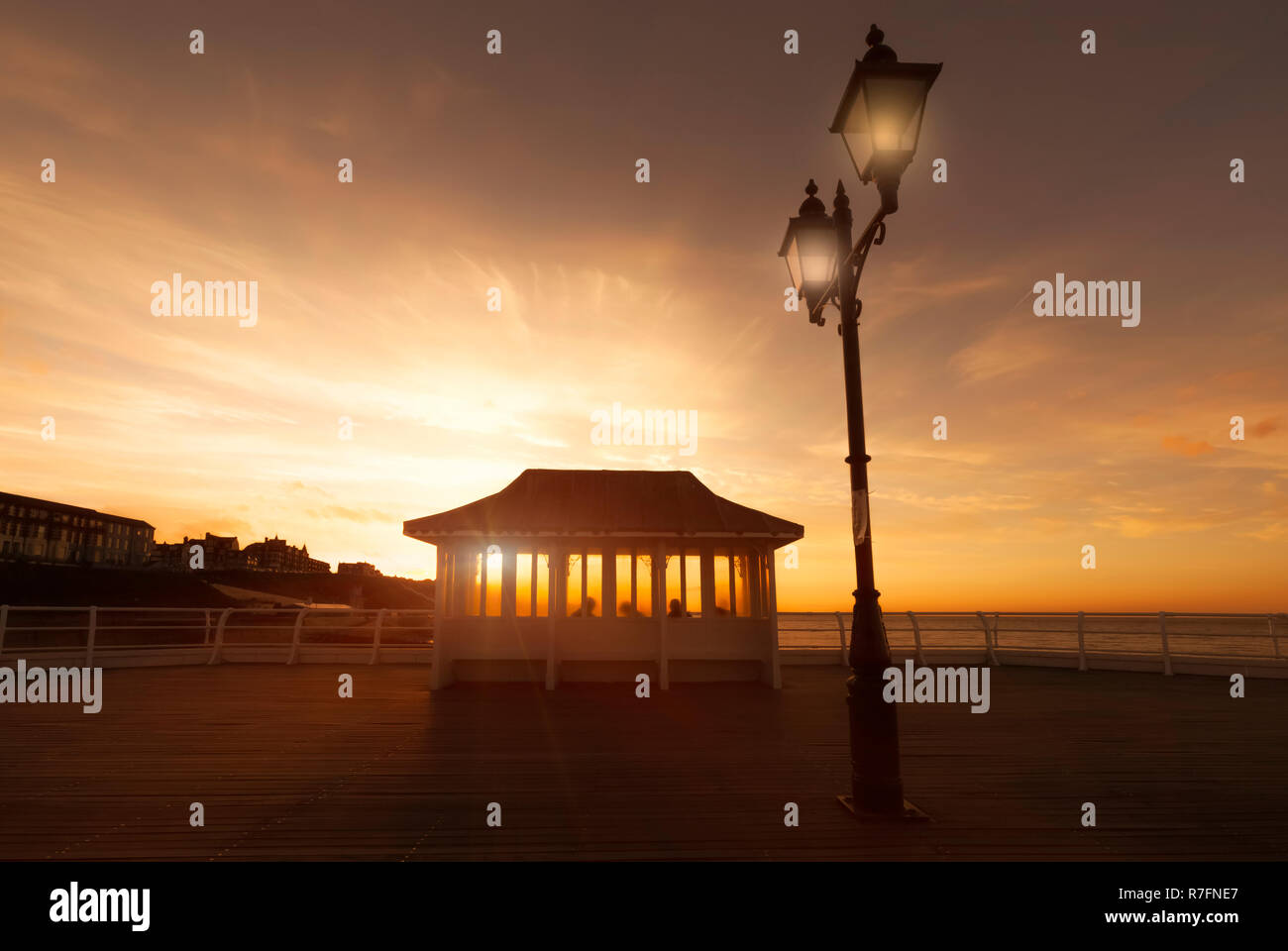 Historic victorian pier shelter at sunset with people in silhouette sat watching the orange glowing sun setting behand the horizon. Old lamp post give Stock Photo