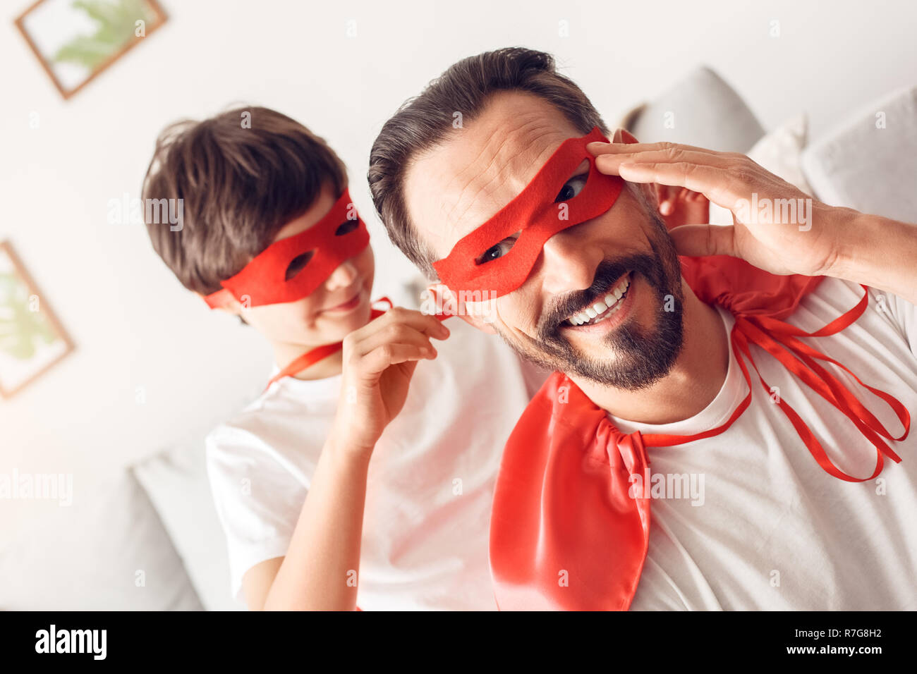 Father and son in superheroe costumes at home boy helping tying mask to dad smiling close-up Stock Photo