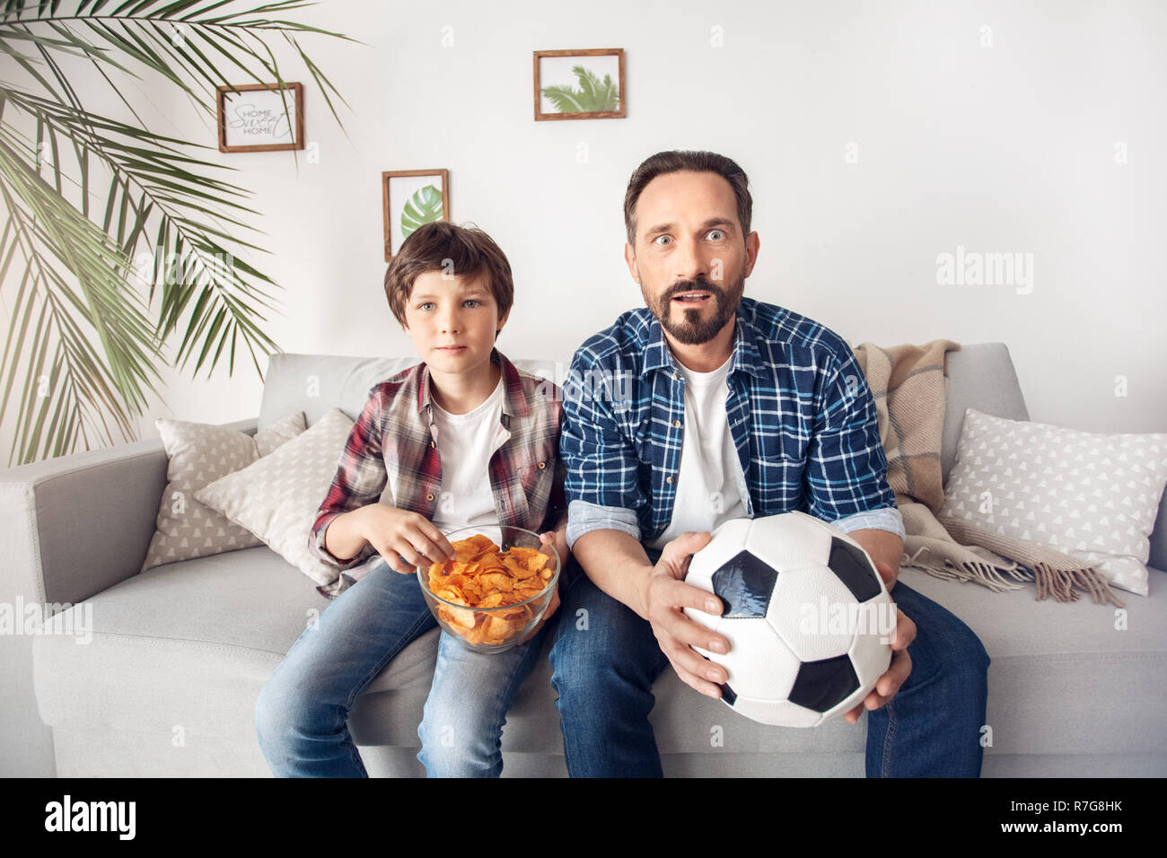Father and little son at home sitting on sofa boy eating potato chip dad with ball watching soccer looking camera impressed Stock Photo