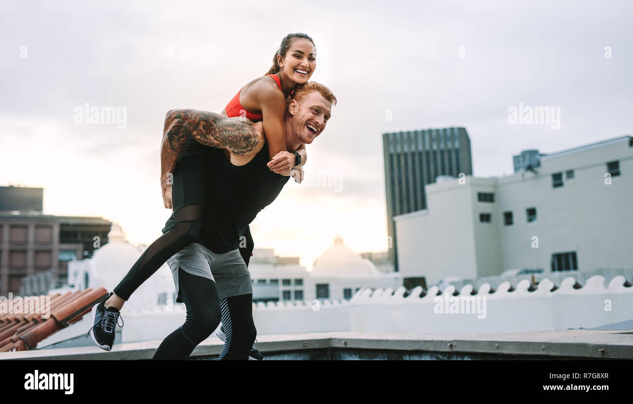 Athlete carrying a fitness woman on his back and walking on rooftop. Smiling fitness woman piggy riding on a man while training on rooftop. Stock Photo