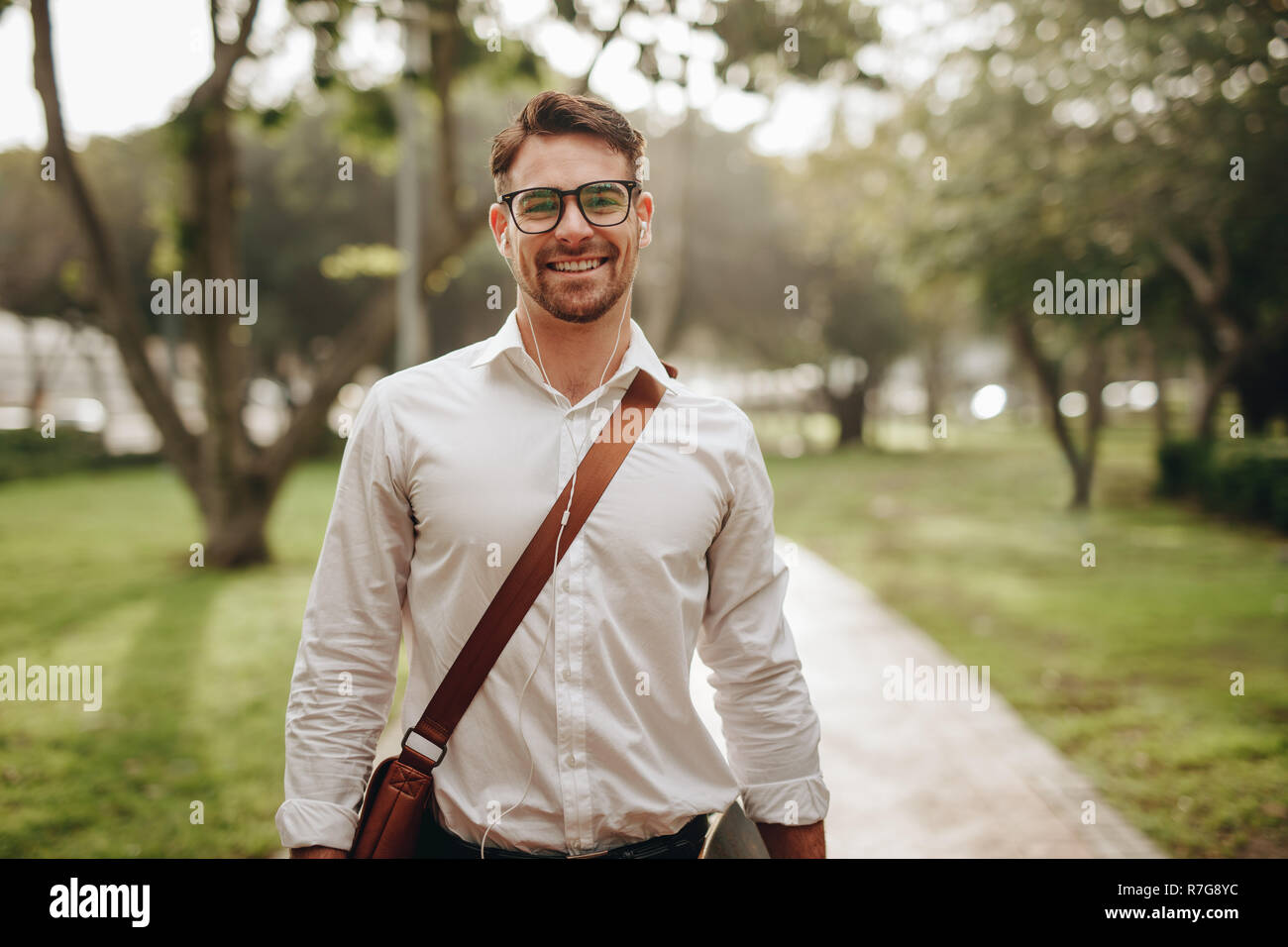 Businessman going to office by walk listening to music on earphones. Happy businessman wearing office bag standing in the street while going to work. Stock Photo