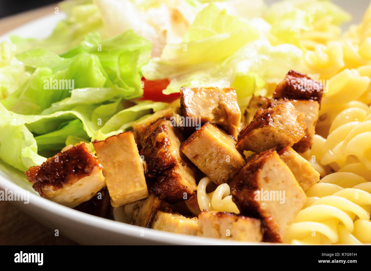 Close up of cooked tofu pieces with pasta and salad in a white bowl on light wood table. Vegetarian and vegan meal. Stock Photo