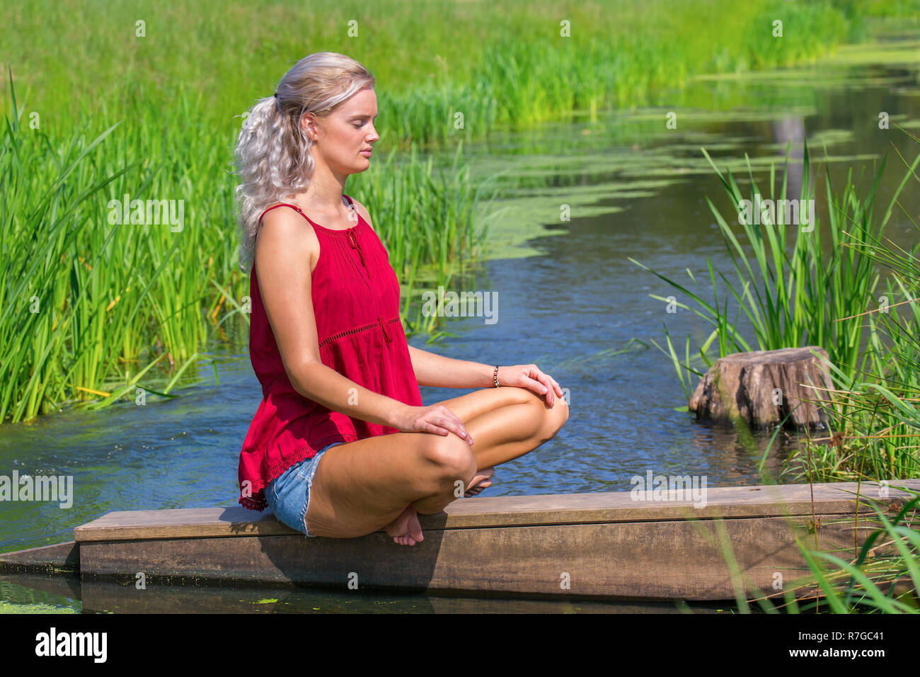 Young blonde caucasian woman meditates at river in nature Stock Photo