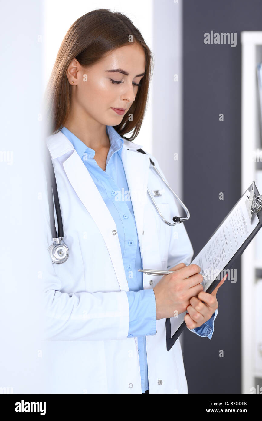 Doctor woman filling up medical form while standing near window in hospital office. Happy physician at work. Medicine and health care concept Stock Photo