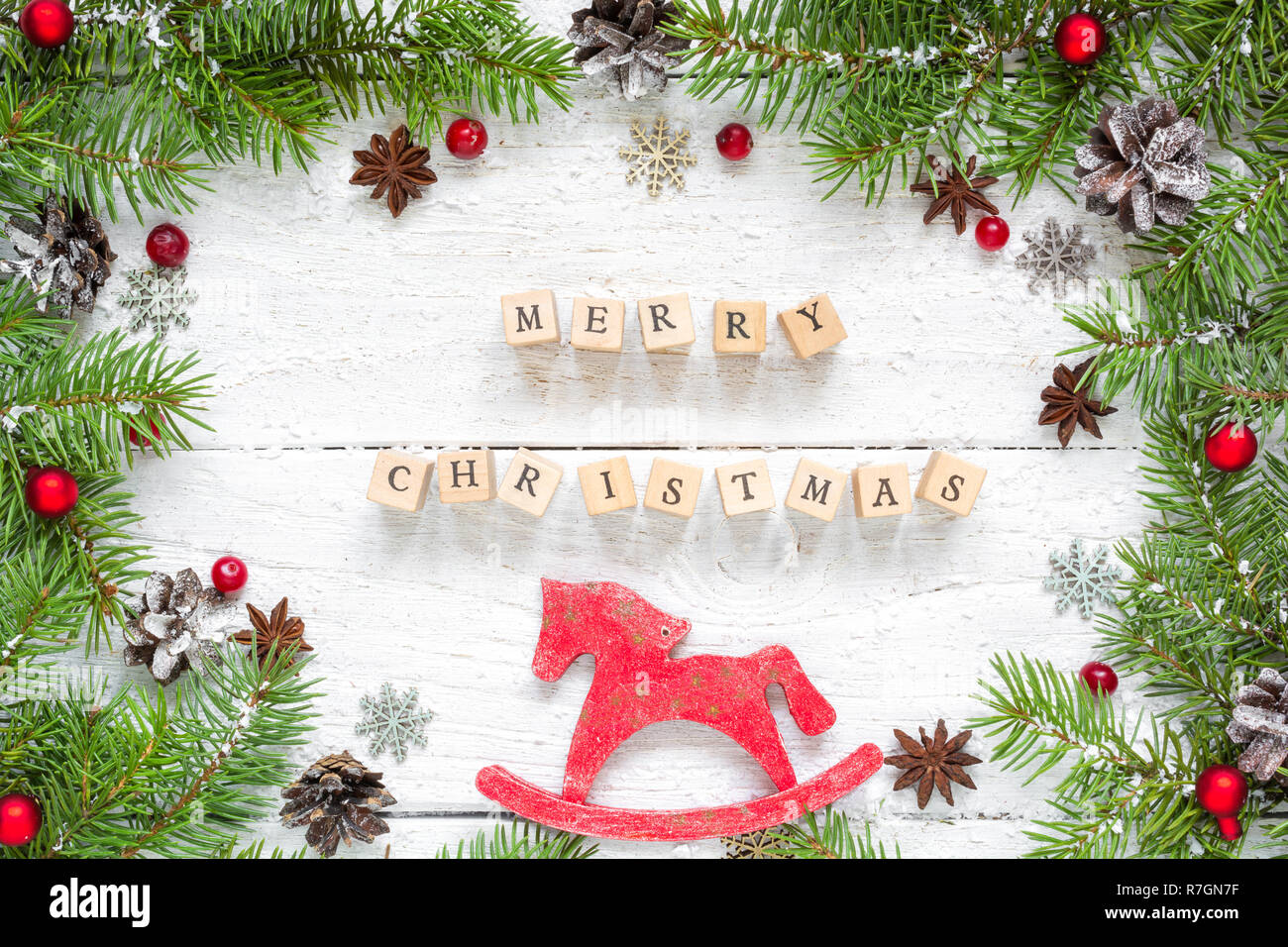 merry christmas inscription in frame made of fir branches, festive decorations, retro toy horse and pine cones on white wooden table. Christmas backgr Stock Photo