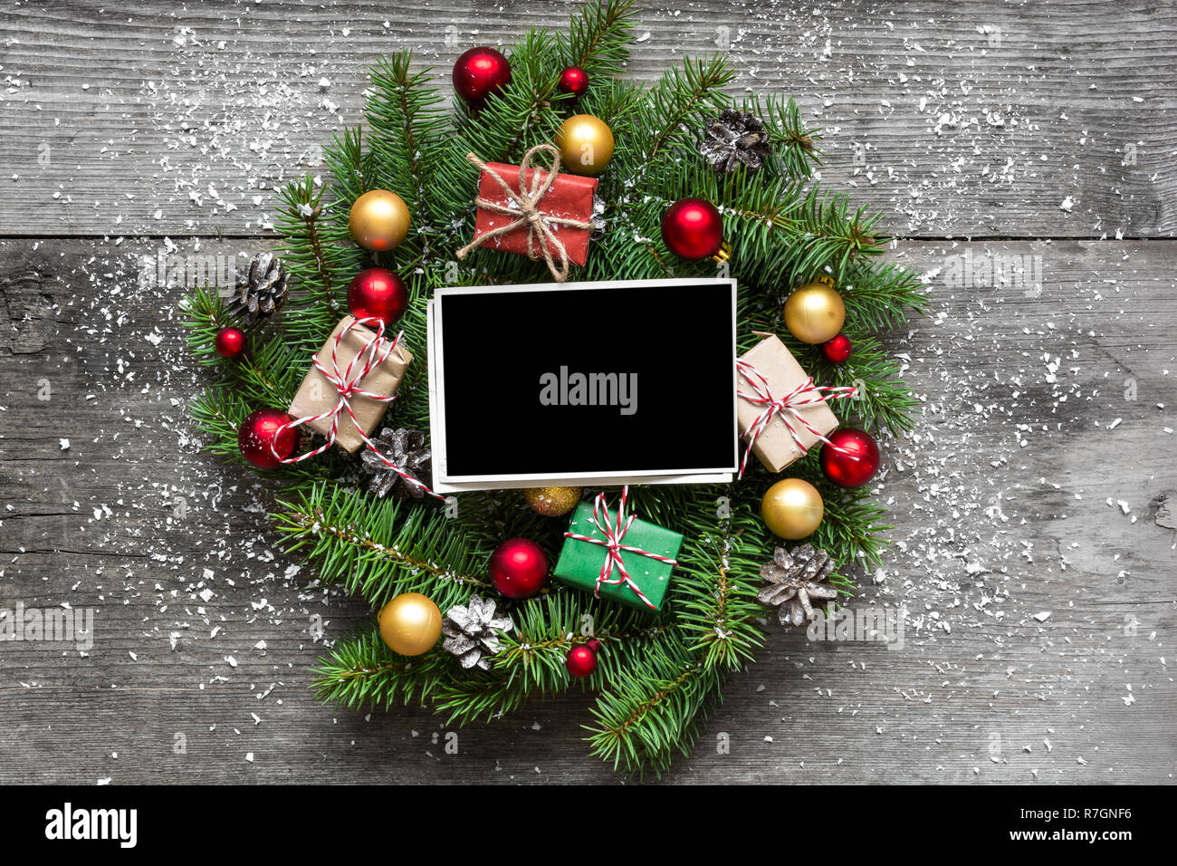 blank photo frame in christmas wreath with decorations and gift boxes on rustic wooden table covered with snow. top view Stock Photo