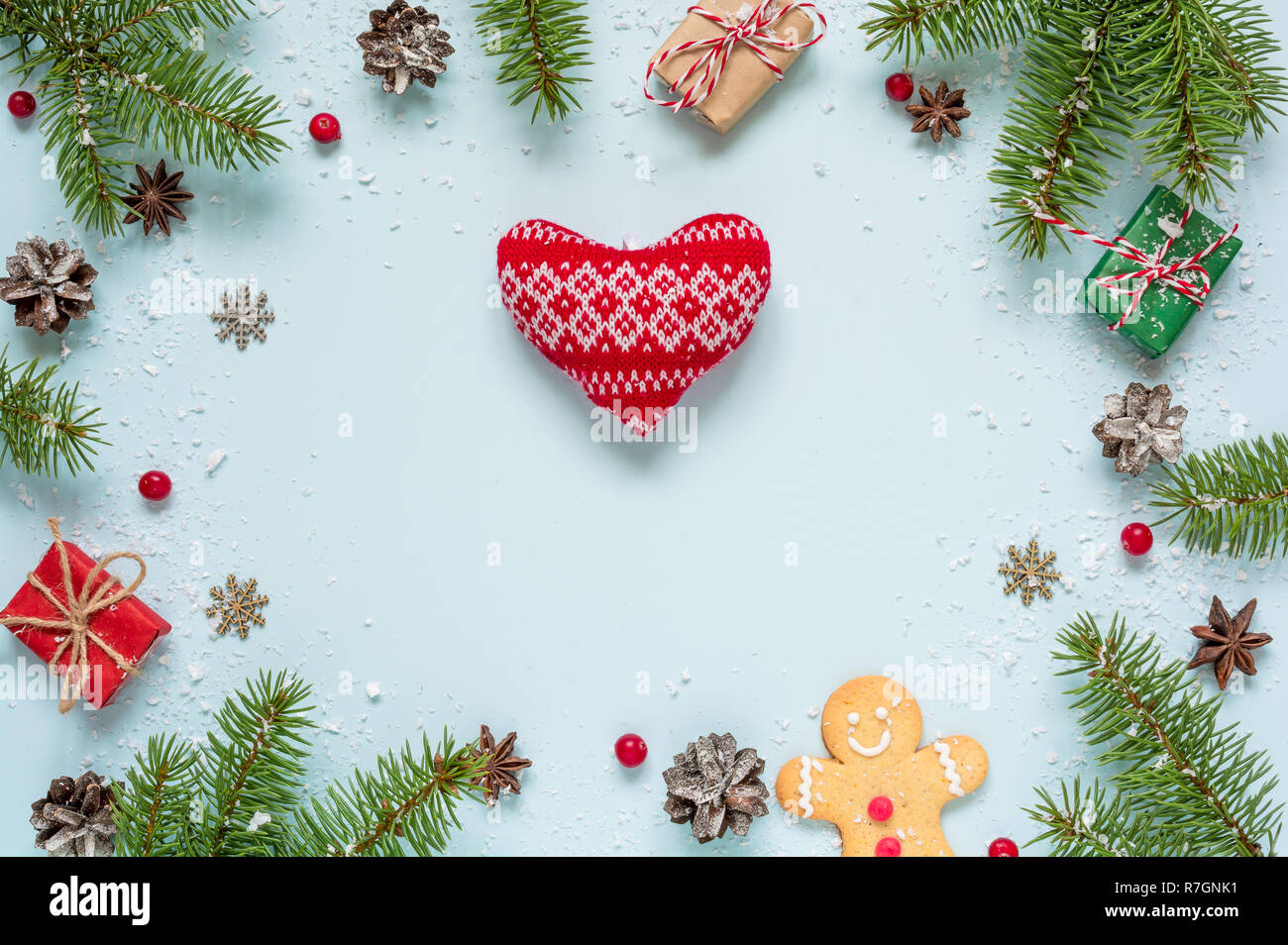 Christmas frame made of fir branches, red berries, gift boxes and pine cones with knitted heart on blue background. Christmas background. Flat lay. to Stock Photo