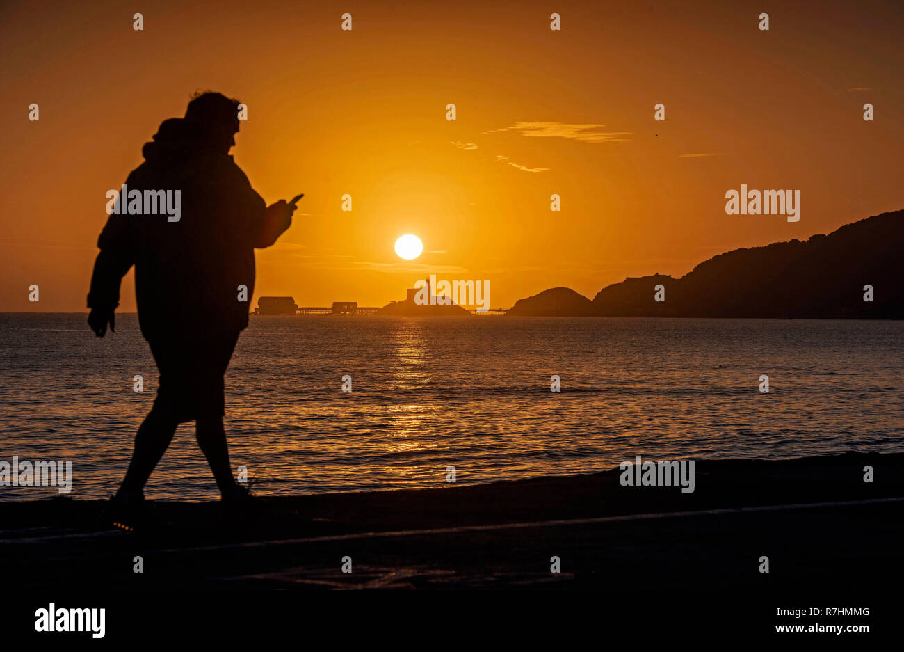 Swansea, UK. 10th Dec, 2018. UK Weather: A walker checks his mobile phone as the sun rises over the Mumbles lighthouse and Pier near Swansea today on the start of a stunning December day. Credit: Phil Rees/Alamy Live News Stock Photo