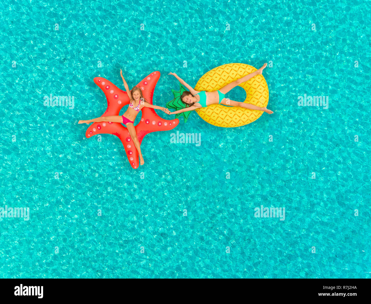 Aerial view of two girls floating on inflatable mattress holding hands on transparent sea. Stock Photo