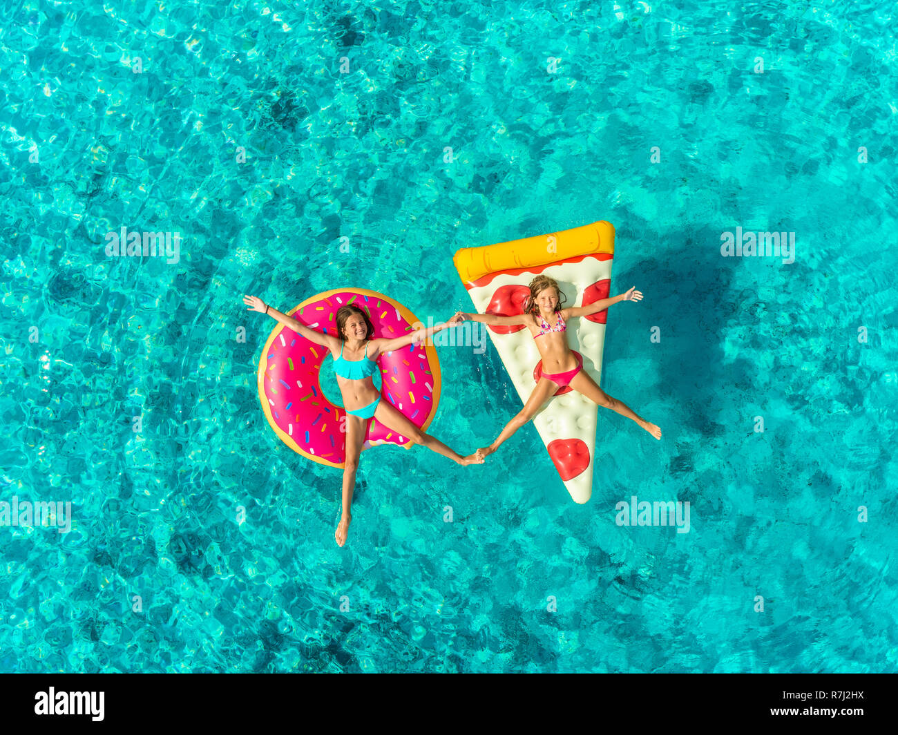 Aerial view of two young girls floating in sea on pizza and donut shaped inflatable holding hands, touching feet and smiling. Stock Photo