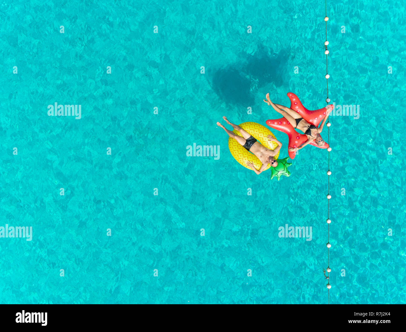 Aerial view of man and woman floating by string buoy on inflatable mattresses in clear blue sea. Stock Photo