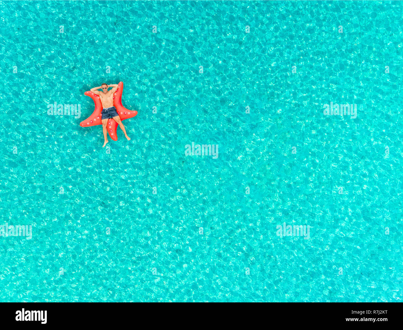 Aerial view of man floating on inflatable star fish mattress with arms folded behind head on transparent sea. Stock Photo
