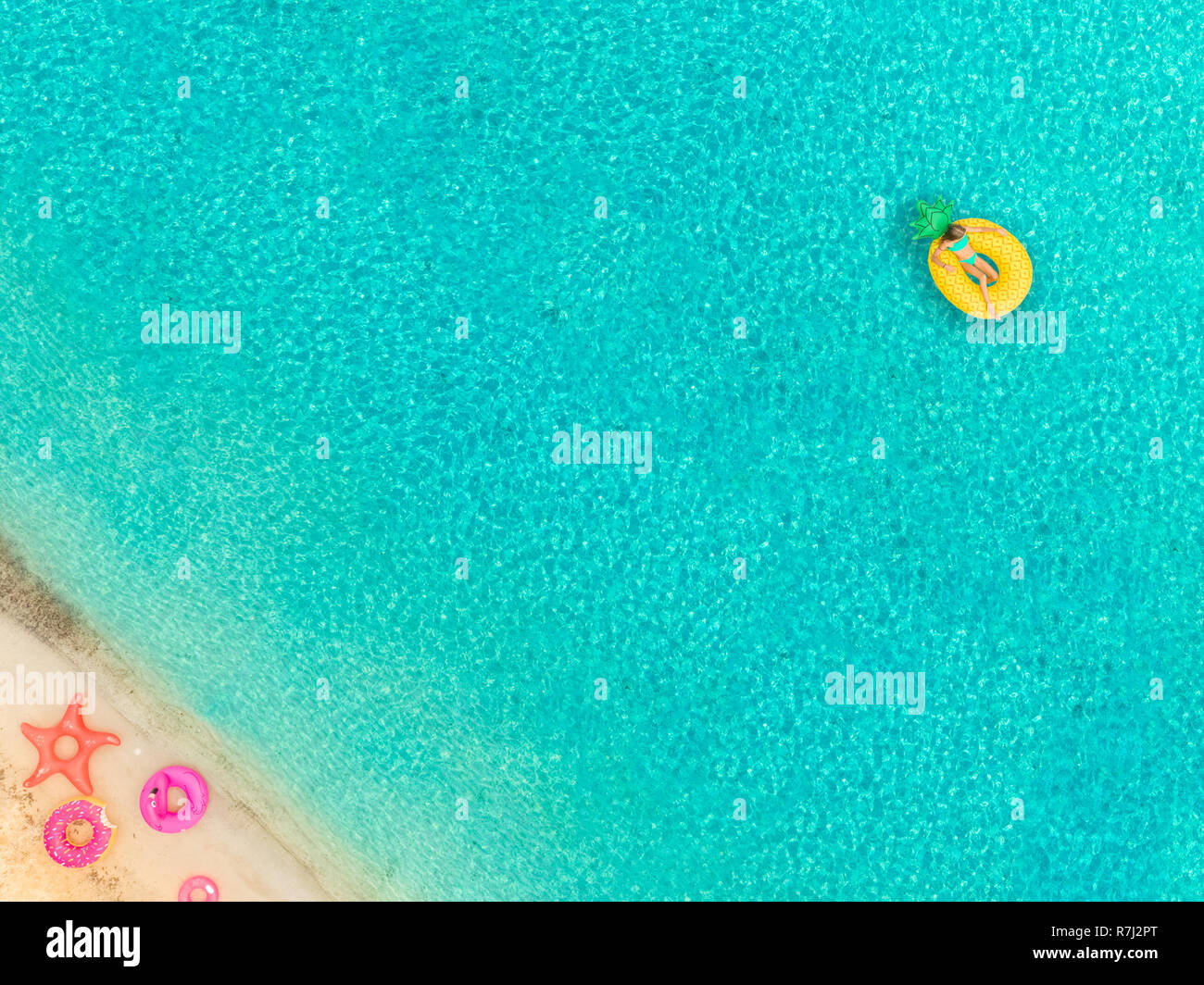 Aerial view of woman floating on inflatable pineapple mattress by sandy beach and inflatable rings. Stock Photo