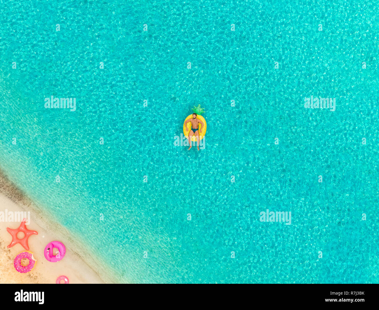 Aerial view of man floating on inflatable pineapple mattress by sandy beach and inflatable rings. Stock Photo