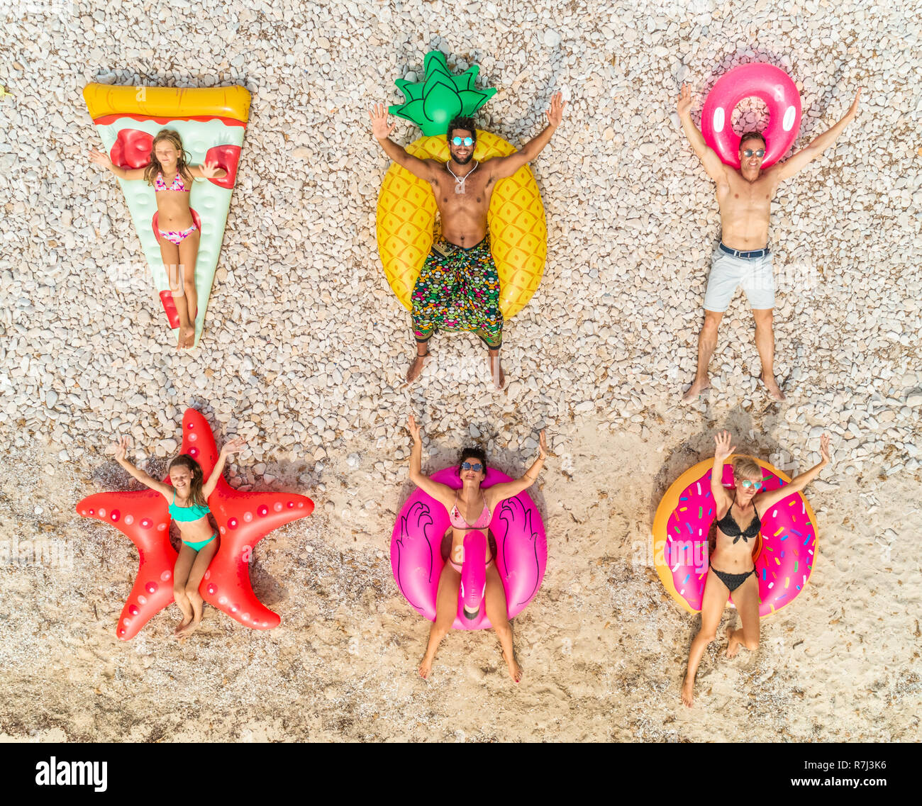 Aerial close up view of friends lying on big inflatable pineapple, pizza, star, donut, flamingo shaped mattresses on beach. Stock Photo