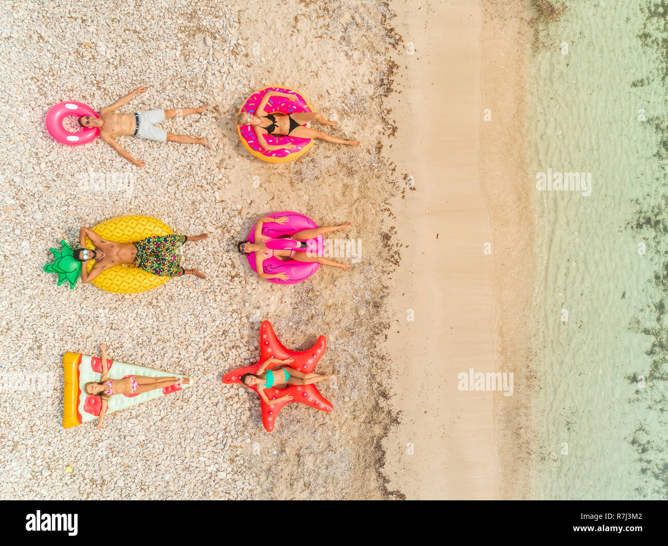 Aerial close up view of people lying on big inflatable pineapple, pizza, star, donut, flamingo shaped mattresses on beach. Stock Photo