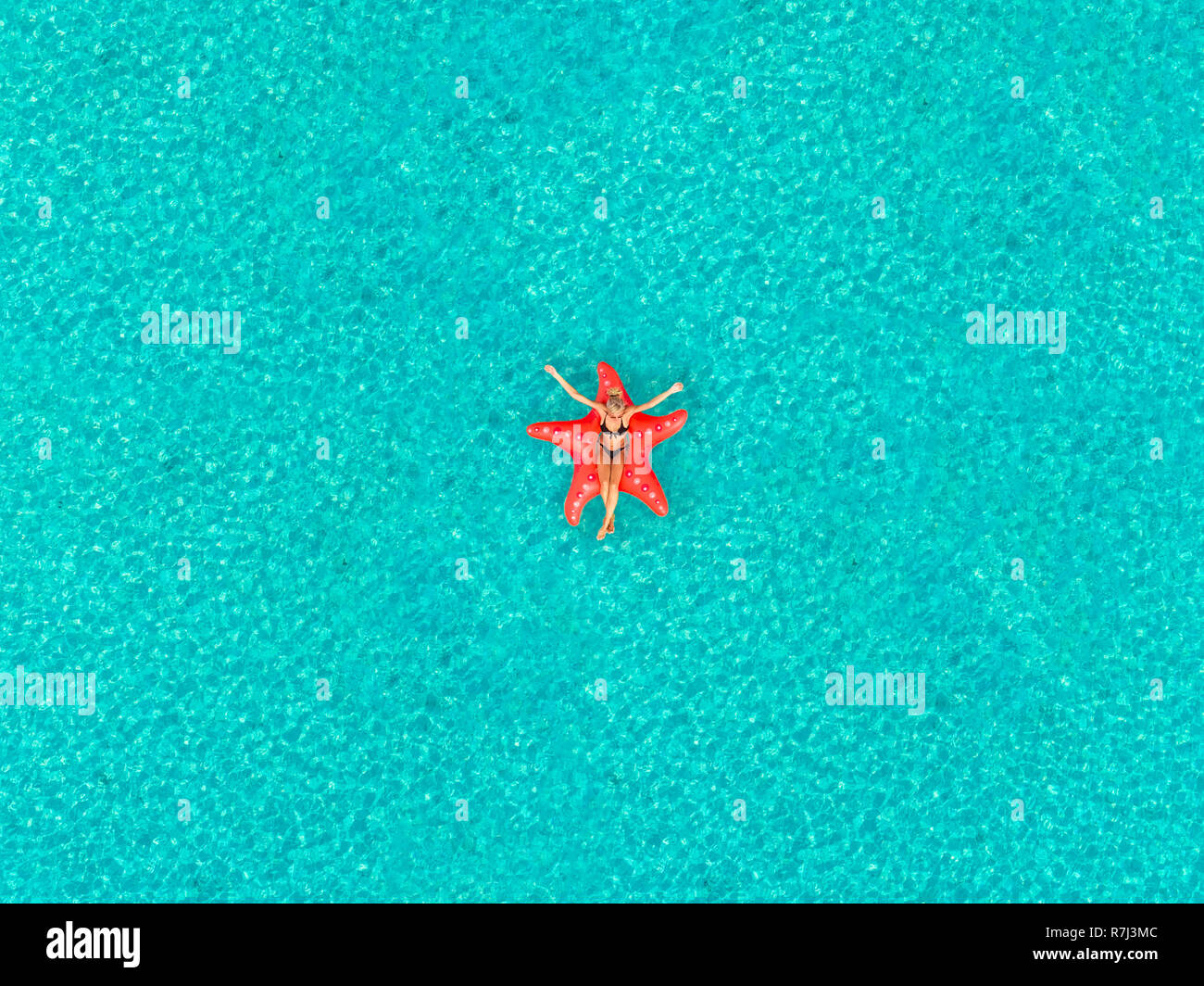 Aerial view of woman floating on inflatable star shaped mattress with arms up in the air on transparent sea. Stock Photo