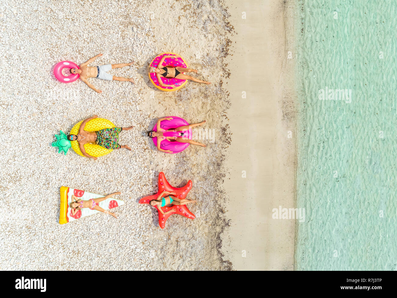 Aerial view of people lying on inflatable pineapple, pizza, star, donut, flamingo shaped mattresses on beach. Stock Photo