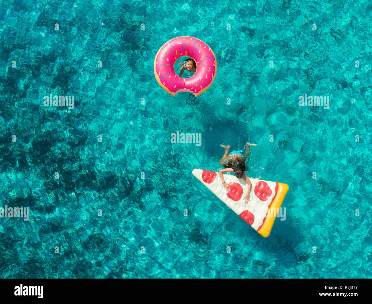 Aerial view of two young girls playing in sea one on pizza, other looking through donut shaped inflatable, smiling. Stock Photo