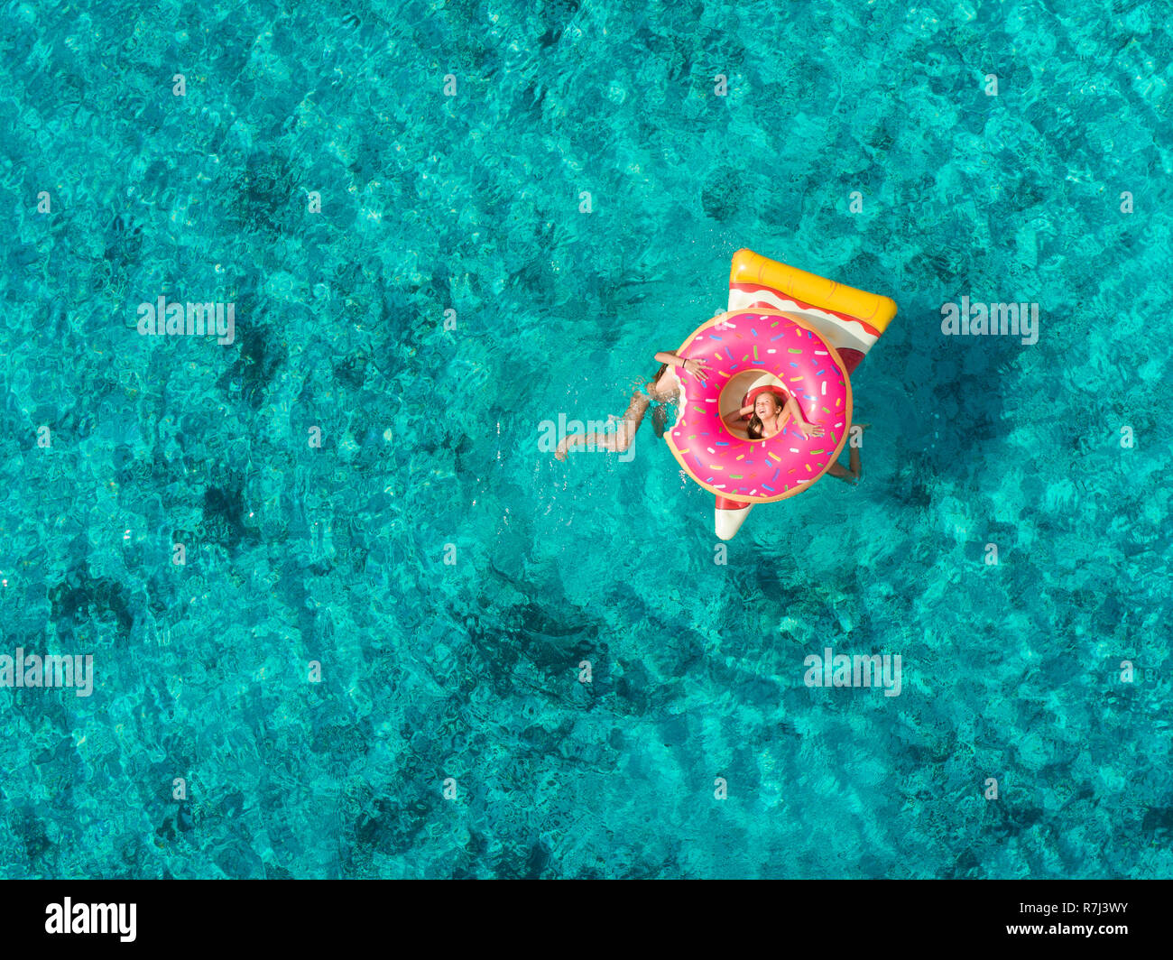 Aerial view of two young girls playing in sea on pizza and donut shaped inflatables. Stock Photo