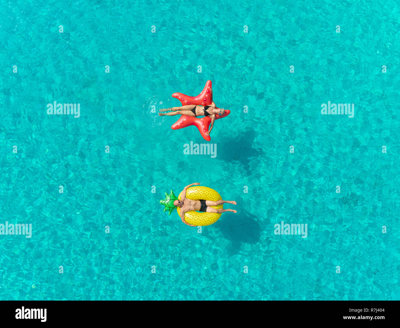 Aerial close up view of couple floating on inflatable star and pineapple shaped mattresses holding hands. Stock Photo