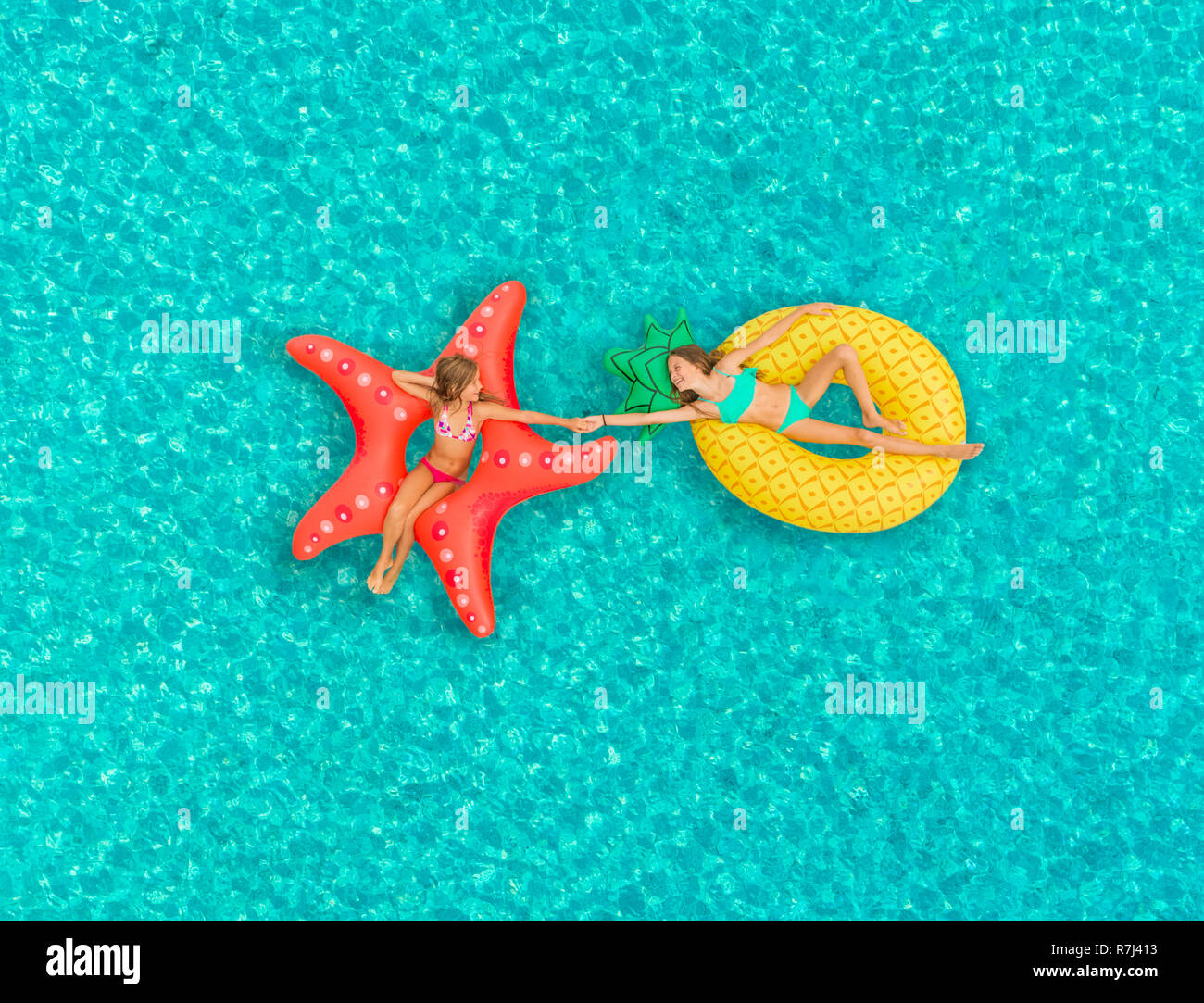 Aerial view of two girls floating on inflatable mattress holding hands on transparent sea. Stock Photo
