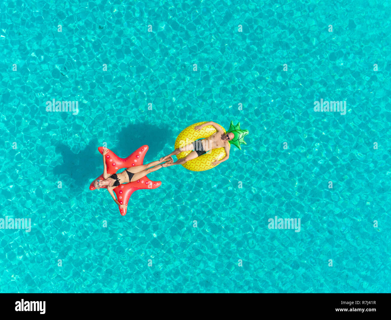 Aerial view of man and woman floating on inflatable mattresses touching each others feet on transparent sea. Stock Photo