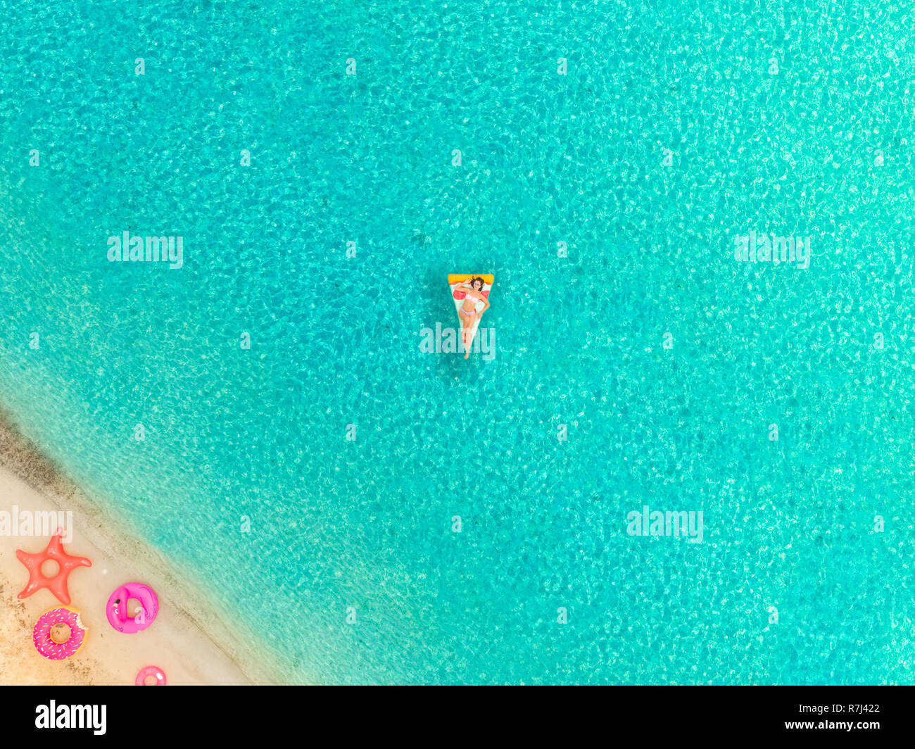 Aerial view of woman floating on inflatable pizza mattress by sandy beach and inflatable rings. Stock Photo