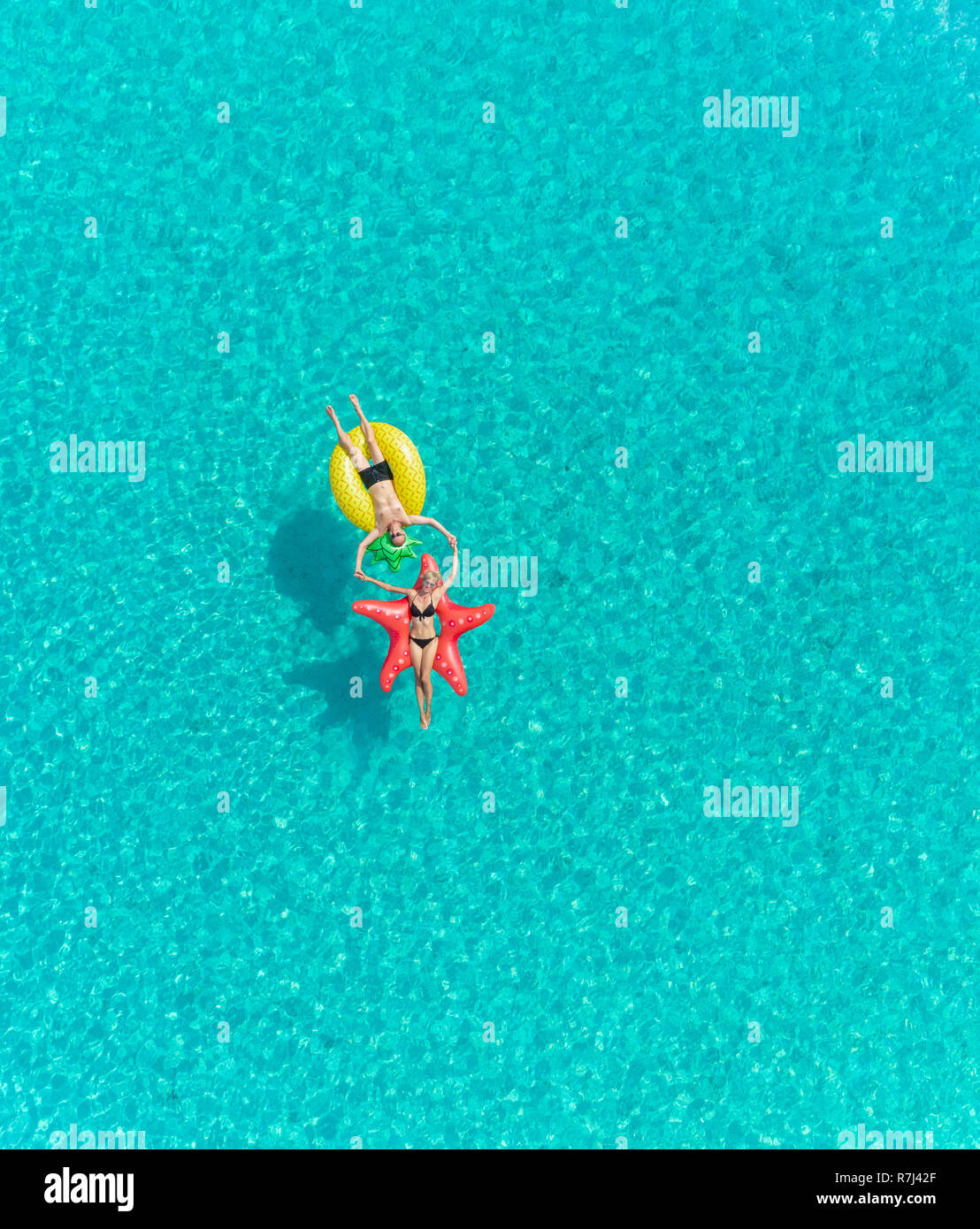 Aerial view of man and woman floating on inflatable star and pineapple shaped mattresses joining hands. Stock Photo