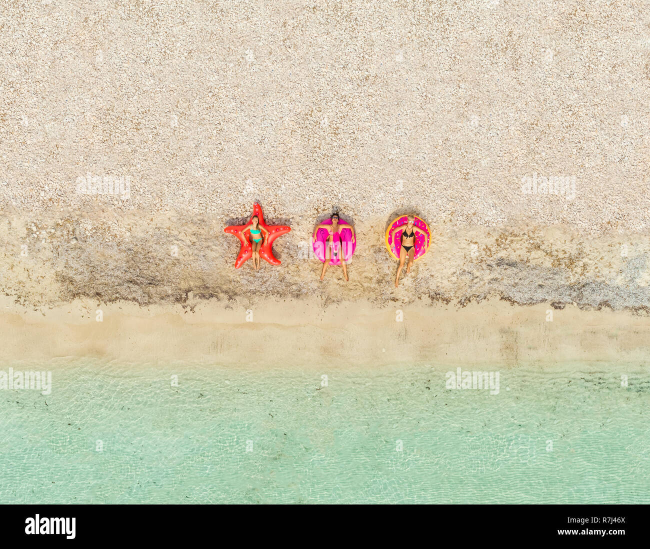 Aerial  view of three women lying on big inflatable star, donut, flamingo shaped mattresses on beach. Stock Photo
