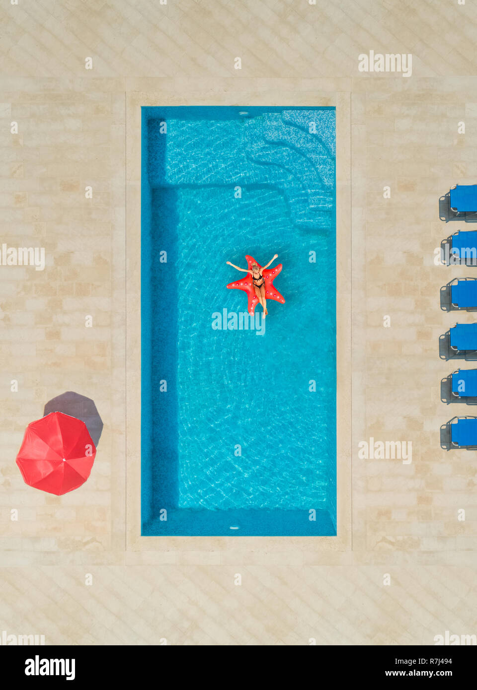 Aerial view of woman on star shapes inflatable mattress in swimming pool surrounded by deck chairs and parasol. Stock Photo