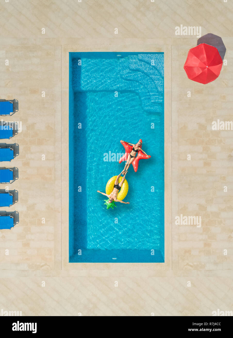 Aerial view of woman and a man on inflatable mattress in swimming pool surrounded by deck chairs and parasol. Stock Photo