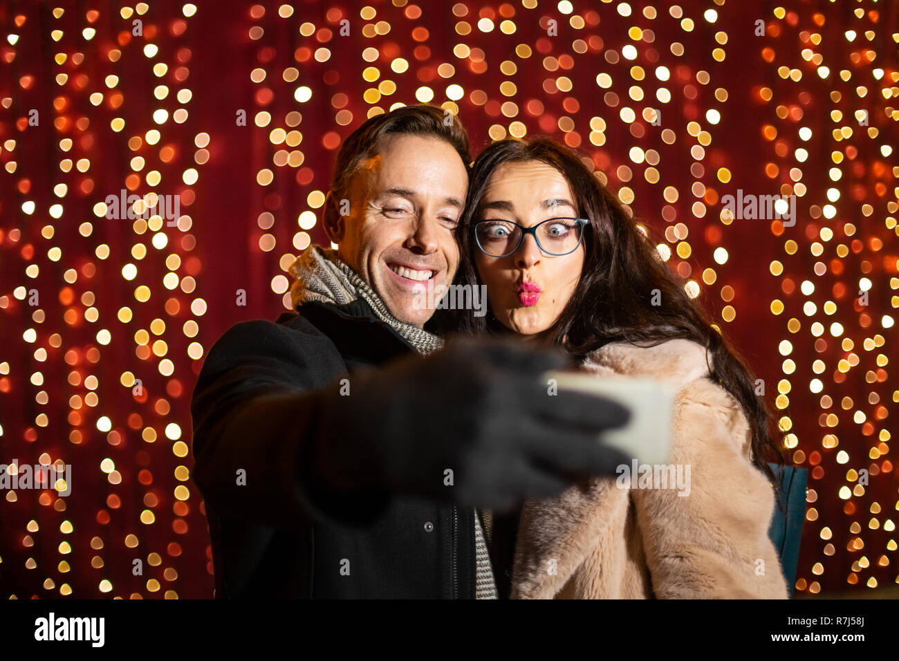 Two friends taking selfie in front of light wall at Christmas market. Stock Photo