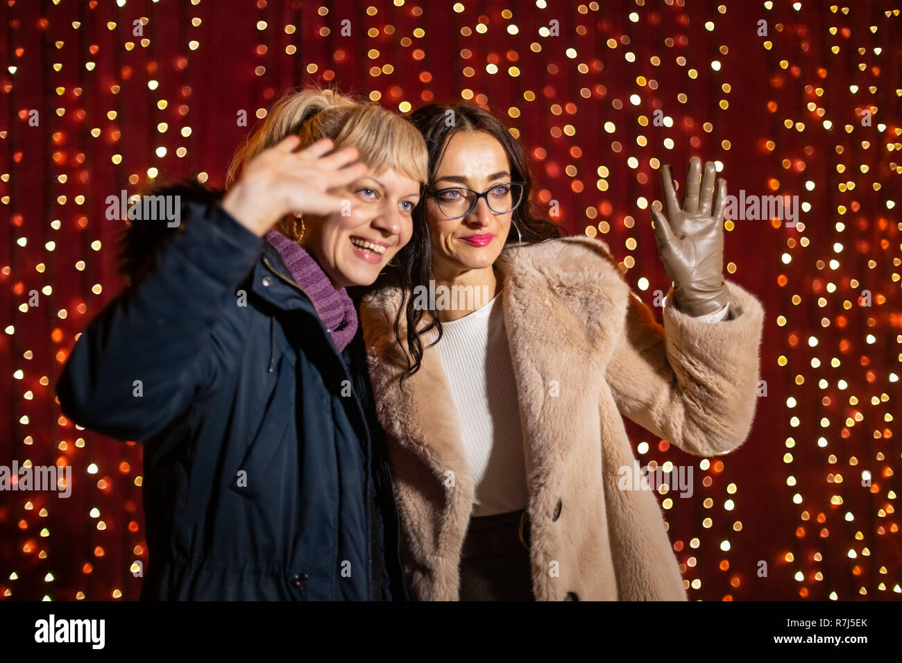Two attractive women posing for photo in front of light wall at Christmas market. Stock Photo