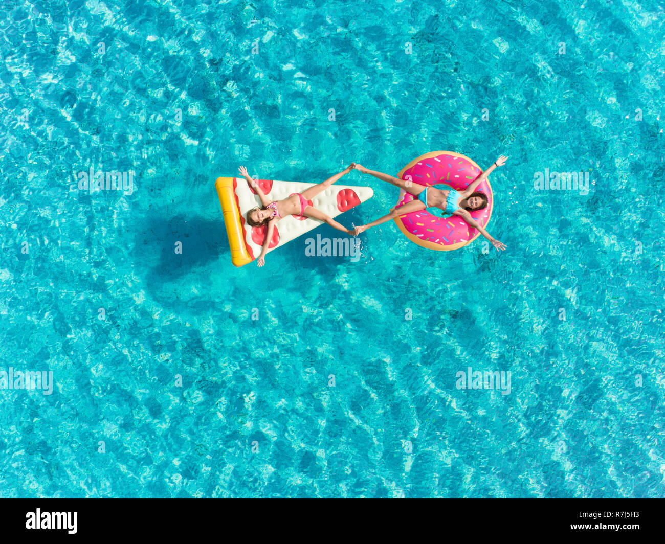 Aerial view of two young girls floating in sea on  pizza and donut shaped inflatables touching feet. Stock Photo