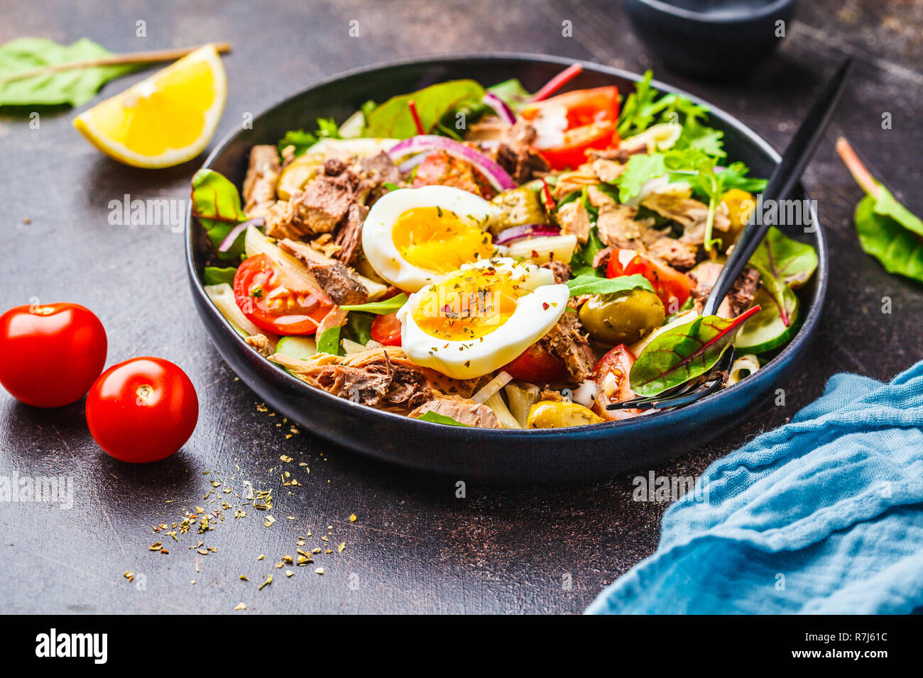 Tuna salad with pasta, olives, vegetables and poached egg in a black plate on a dark background. Stock Photo