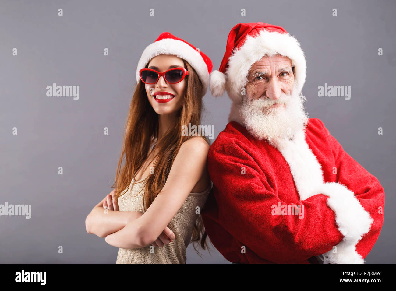 Santa Claus and young mrs. Claus wearing Santa hat and sunglasses standing and smiling on the gray background New Year Christmas holidays souvenirs gifts shopping discounts shops Snow Maiden Santa Cla...