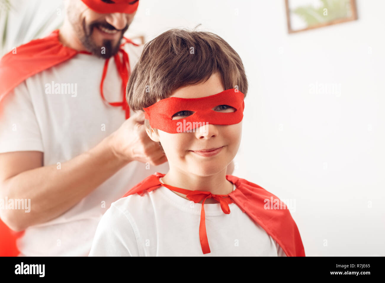 Father and son in superheroe costumes at home standing boy smiling happy close-up while man tying knot on his mask Stock Photo
