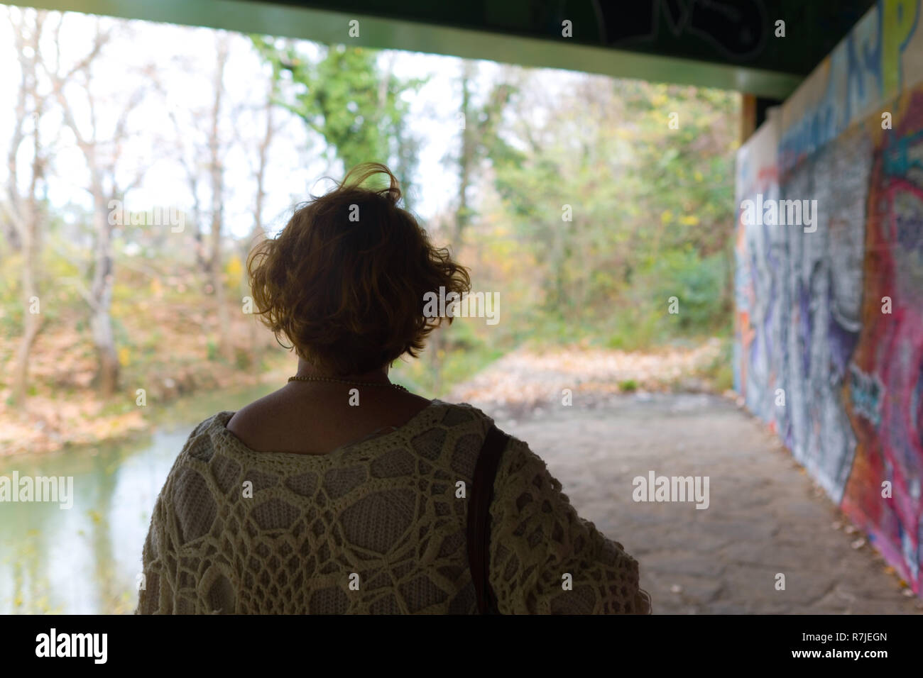 cropped rear view of middle aged woman shoulders and head walking under graffitied bridge by river Stock Photo