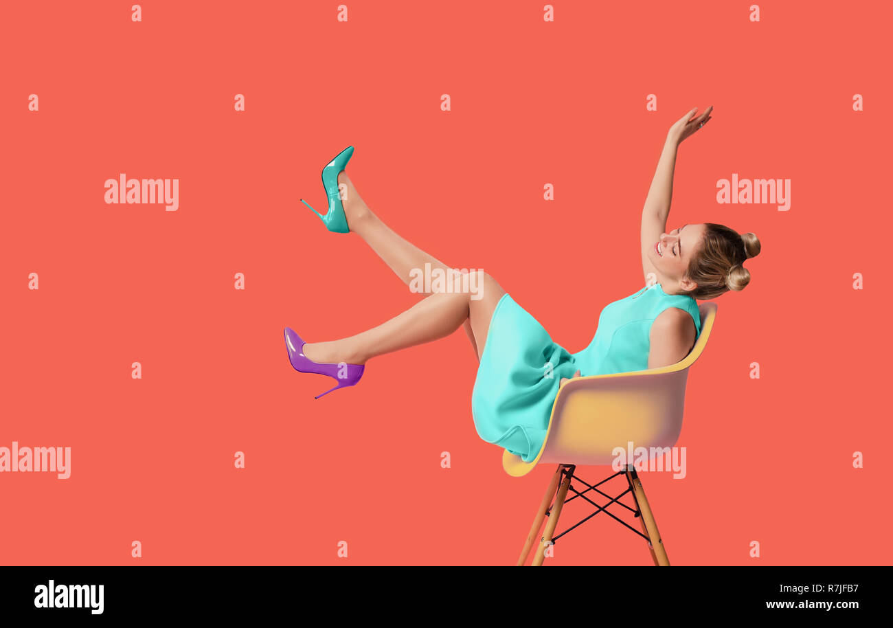 trendy young woman wearing turquoise dress and modern high heels shoes sitting on chair with raised legs, smiling , against living coral background Stock Photo