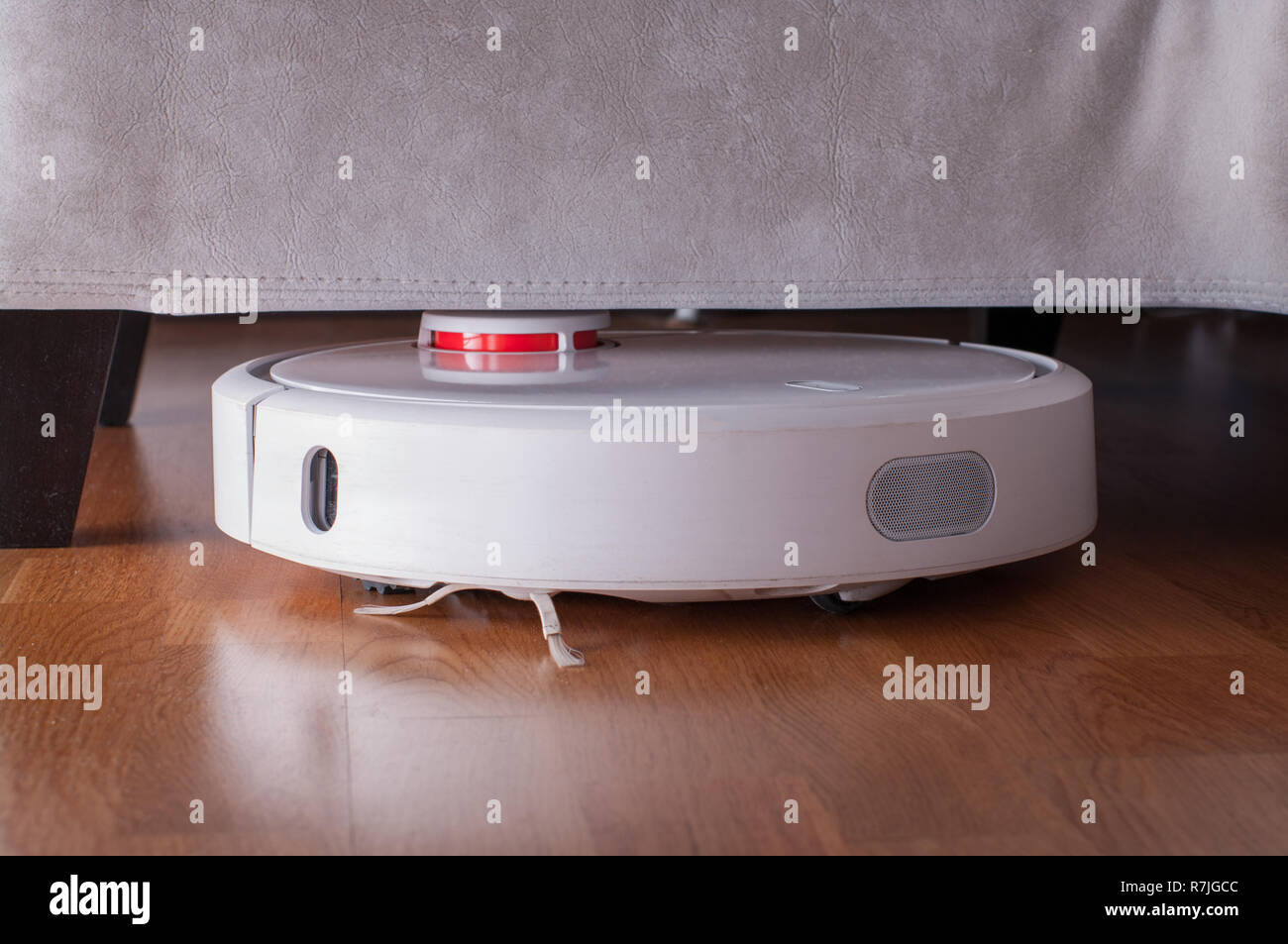 Robotic vacuum cleaner runs under sofa in room on laminate floor. Robot