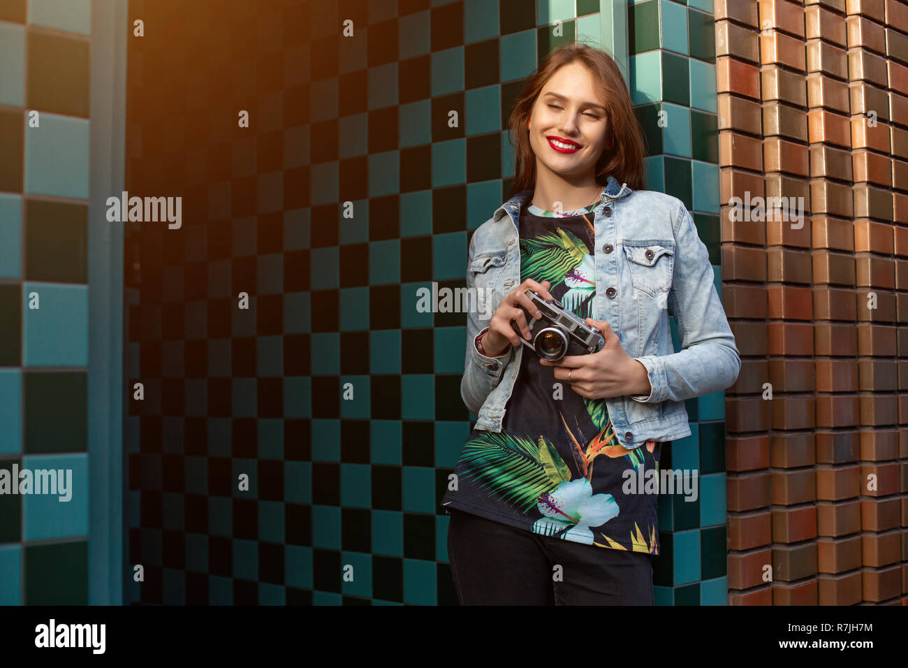 Cool funny girl model with retro film camera wearing a denim jacket, dark hair outdoors over city wall in a cage background. Sun flare Stock Photo