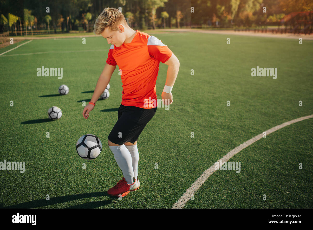 Young football player excercising on lawn. He kicks ball with leg. Guy is concentrated. Sunny summer weater is outside. Stock Photo