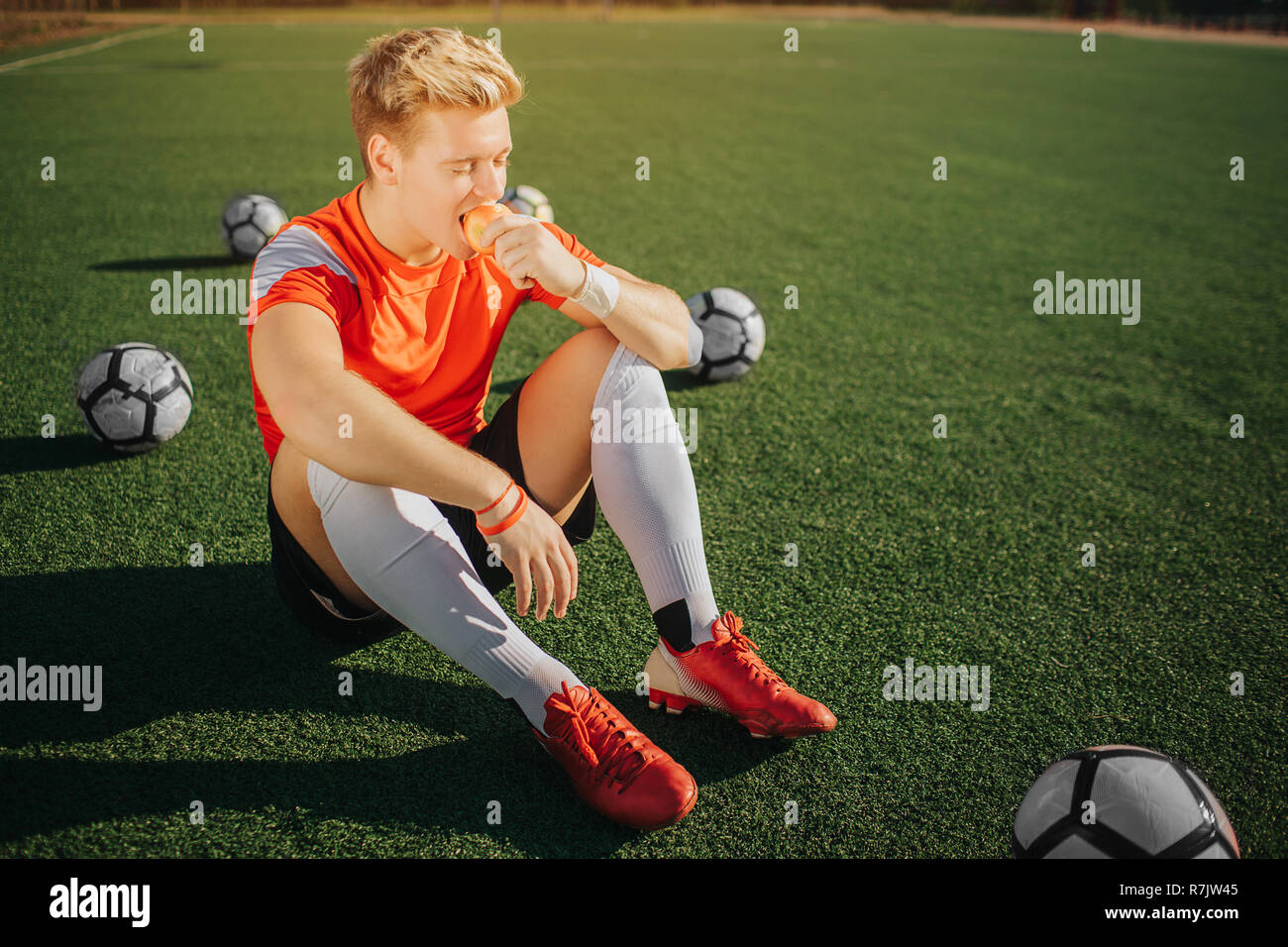 Hungry young player sitting on lawn and eating apple. He has rest. Three balls are behind him. Fourth is in front. Sunny weather is outside. Stock Photo